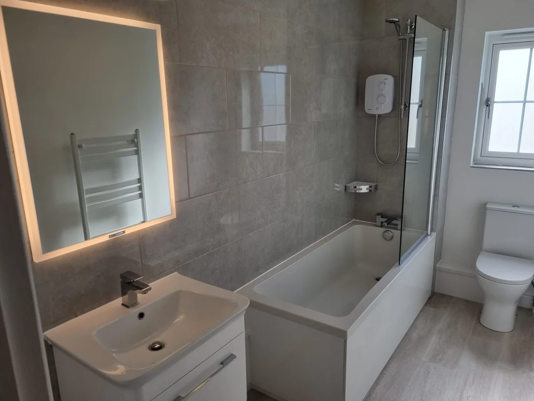 Bathroom fitted by Pete: bath with a glass shower screen, grey tiled wall, illuminated mirror and a white vanity basin